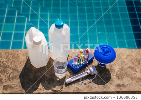 Pool water test kit, floating chemical dispenser, thermometer, bottles, and measuring cup on swimming pool edge 136686355