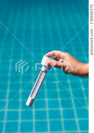 Hand holding pool thermometer above clear blue swimming pool water, checking temperature during routine pool maintenance and water quality control 136686376