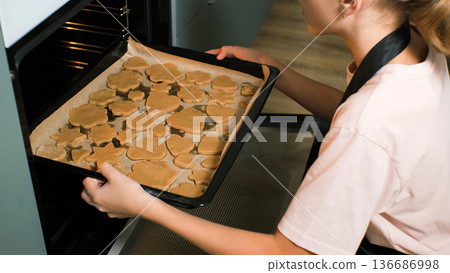 A teenage girl wearing an apron places a tray of raw ginger cookies into the oven, baking homemade bakery A teenage girl wearing an apron places a tray of raw ginger cookies into the oven, baking homemade bakery 136686998