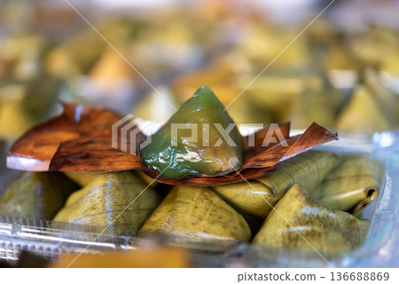 Traditional Thai sweet dessert wrapped in banana leaf with green jelly texture displayed in market tray authentic local food close up macro with soft background bokeh Traditional Thai sweet dessert wrapped in banana leaf with green jelly texture displayed in market tray authentic local food close up macro with soft background bokeh 136688869