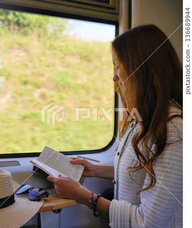 Woman sitting by train window with book and hat on table 136689944