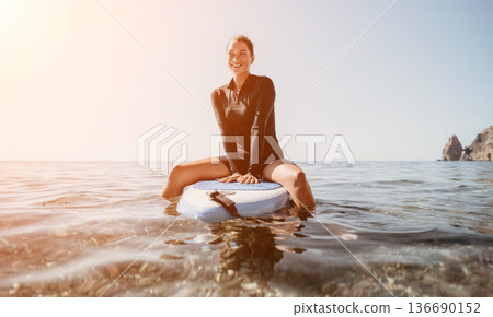 Woman Surfer Beach Sunlight - A woman in a wetsuit sits on a surfboard in the ocean, looking out at the sun setting on the horizon. 136690152