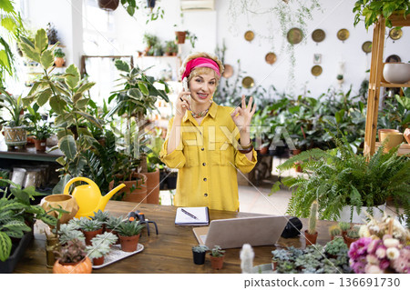 A woman in a yellow shirt gestures okay while talking on the phone in her plant shop A woman in a yellow shirt gestures okay while talking on the phone in her plant shop 136691730