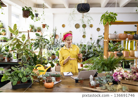 A woman in a yellow shirt is showing a cactus to the camera in a plant shop, surrounded by greenery and a laptop 136691746