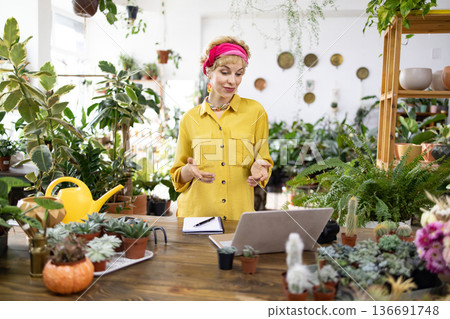 A woman in a yellow shirt gestures while on a video call in a plant shop surrounded by greenery 136691748
