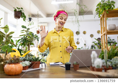 A woman waves while on a video call in her plant shop, surrounded by lush greenery and succulents A woman waves while on a video call in her plant shop, surrounded by lush greenery and succulents 136691759