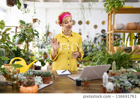 A woman in a yellow shirt waves during a video call in a plant-filled shop A woman in a yellow shirt waves during a video call in a plant-filled shop 136691761