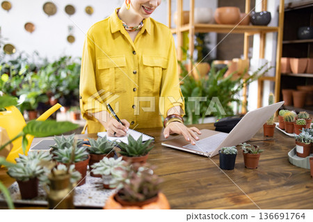 A woman in a yellow shirt works on a laptop and takes notes surrounded by plants and succulents A woman in a yellow shirt works on a laptop and takes notes surrounded by plants and succulents 136691764