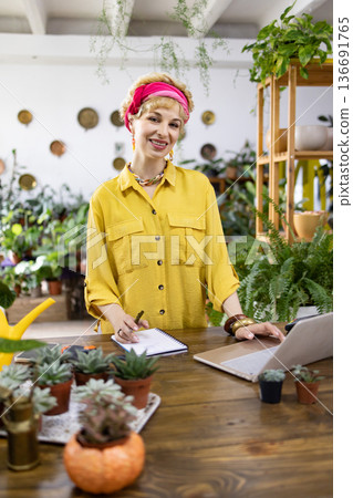 A smiling woman works at a desk surrounded by plants, succulents, and a laptop, creating a welcoming atmosphere A smiling woman works at a desk surrounded by plants, succulents, and a laptop, creating a welcoming atmosphere 136691765