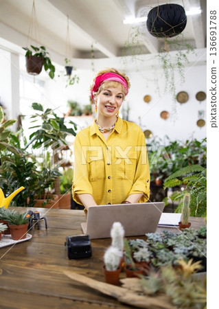 A smiling woman works on a laptop surrounded by lush plants in a bright, inviting shop A smiling woman works on a laptop surrounded by lush plants in a bright, inviting shop 136691788