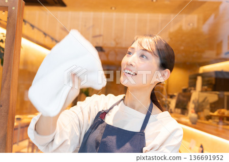 一位年輕的女店員穿著圍裙,正在擦拭咖啡館的窗戶。 一位年輕的女店員穿著圍裙,正在擦拭咖啡館的窗戶。 136691952