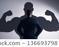 A confident businessman in a suit stands tall, his shadow revealing a muscular physique, symbolizing inner strength and ambition in a competitive corporate world. 136693798