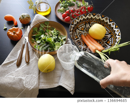 Anonymous person pours water from bottle into glass. Multicolored ceramic plates and patterned bowl with vegetables and herbs on black background. Food and healthy eating. View from above. Anonymous person pours water from bottle into glass. Multicolored ceramic plates and patterned bowl with vegetables and herbs on black background. Food and healthy eating. View from above. 136694685