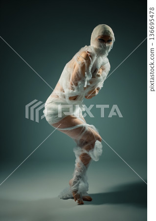 Conceptual portrait of a person wrapped like a mummy in white bandages, struggling in a constrained pose. Conceptual portrait of a person wrapped like a mummy in white bandages, struggling in a constrained pose. 136695478