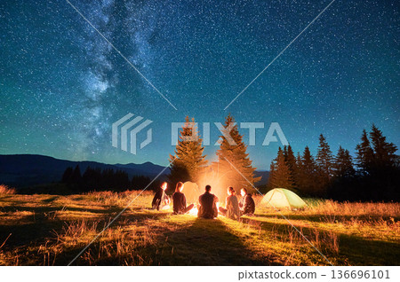 Group of friends hikers resting around campfire, surrounded by illuminated tents under stunning starry sky. Milky Way stretches above, creating magical atmosphere in peaceful mountain wilderness. Group of friends hikers resting around campfire, surrounded by illuminated tents under stunning starry sky. Milky Way stretches above, creating magical atmosphere in peaceful mountain wilderness. 136696101