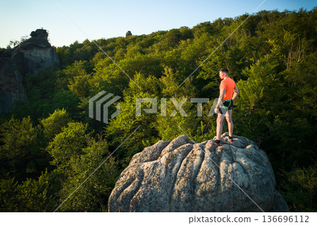 Aerial view of climber standing atop rock formation at Dovbush Rocks in Carpathian mountains, Ukraine. Sun sets, casting warm glow over lush, green forest, distant hills, creating breathtaking view. Aerial view of climber standing atop rock formation at Dovbush Rocks in Carpathian mountains, Ukraine. Sun sets, casting warm glow over lush, green forest, distant hills, creating breathtaking view. 136696112