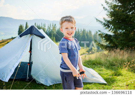 Smiling young boy stands in front of white tent, wearing blue shirt and shorts, holding camping tool. Child enjoys sunny day in picturesque meadow with lush trees and mountains in background. 136696118
