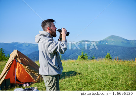 Man stands near orange tent, using binoculars to observe distant mountains. Grassy hilltop, under clear blue sky, lush green forests and rolling hills. Camping gear scattered around tent. 136696120