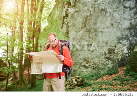 Bearded tourist man studies map while stands in front of large rock in dense forest. Traveler with grey backpack and glasses, planning outdoor adventure, hiking or climbing route. 136696122