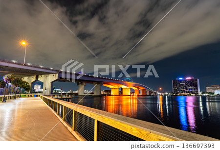 The Acosta Bridge spans the St. Johns River in Jacksonville, Florida. The illuminated bridge and city skyline are viewed from the wooden boardwalk of the Northbank Riverwalk at night. 136697393