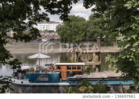 Framed by lush green foliage, a charming houseboat sits moored on the calm waters of the Thames near Kew Bridge. Its upper deck, complete with a white patio umbrella and an array of potted plants. Framed by lush green foliage, a charming houseboat sits moored on the calm waters of the Thames near Kew Bridge. Its upper deck, complete with a white patio umbrella and an array of potted plants. 136697661