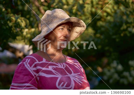 a senior woman in pink dress and straw hat enjoying golden hour sunset in the garden 136698100