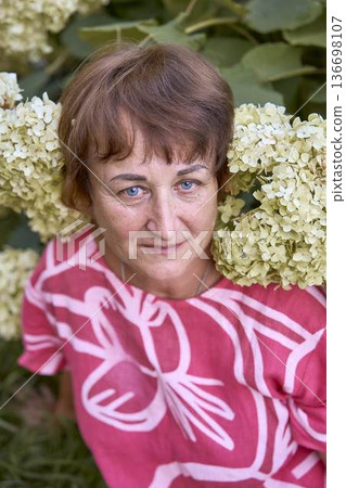 a senior woman sitting among blooming hydrangea flowers in a summer garden 136698107