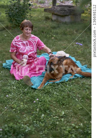 a Senior woman relaxing on a picnic blanket with german shepherd dog and coffee in a garden 136698112