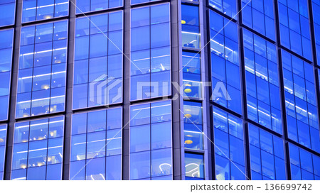 Big glowing windows in modern office building at night, in rows of windows light shines. Fragment of the glass facade of a modern corporate building at night. Modern glass office  in city. 136699742