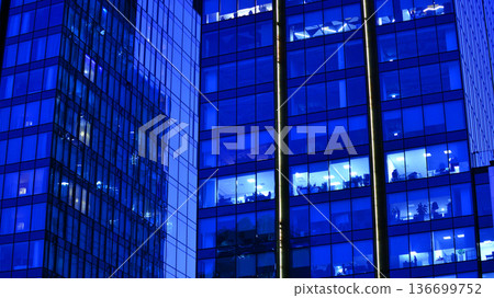 Big glowing windows in modern office building at night, in rows of windows light shines. Fragment of the glass facade of a modern corporate building at night. Modern glass office in city. Big glowing windows in modern office building at night, in rows of windows light shines. Fragment of the glass facade of a modern corporate building at night. Modern glass office in city. 136699752