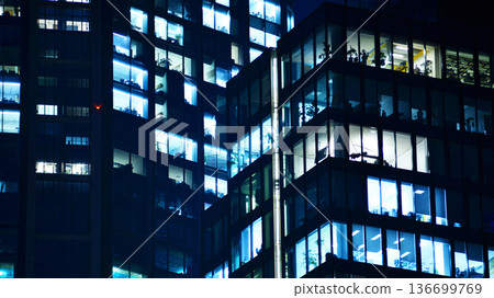 Big glowing windows in modern office building at night, in rows of windows light shines. Fragment of the glass facade of a modern corporate building at night. Modern glass office  in city. 136699769