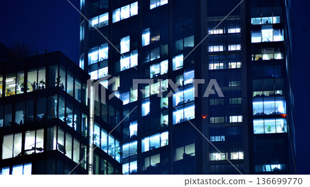 Big glowing windows in modern office building at night, in rows of windows light shines. Fragment of the glass facade of a modern corporate building at night. Modern glass office  in city. 136699770