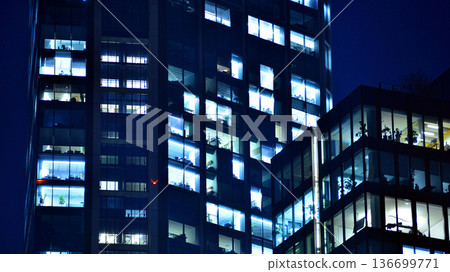 Big glowing windows in modern office building at night, in rows of windows light shines. Fragment of the glass facade of a modern corporate building at night. Modern glass office  in city. 136699771