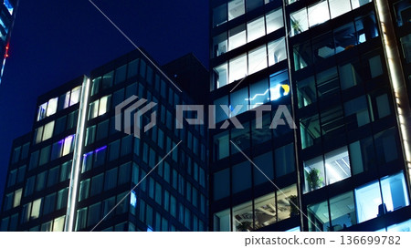 Big glowing windows in modern office building at night, in rows of windows light shines. Fragment of the glass facade of a modern corporate building at night. Modern glass office in city. Big glowing windows in modern office building at night, in rows of windows light shines. Fragment of the glass facade of a modern corporate building at night. Modern glass office in city. 136699782