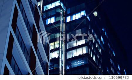 Big glowing windows in modern office building at night, in rows of windows light shines. Fragment of the glass facade of a modern corporate building at night. Modern glass office in city. Big glowing windows in modern office building at night, in rows of windows light shines. Fragment of the glass facade of a modern corporate building at night. Modern glass office in city. 136699783