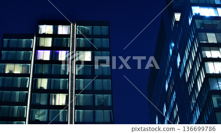 Big glowing windows in modern office building at night, in rows of windows light shines. Fragment of the glass facade of a modern corporate building at night. Modern glass office  in city. 136699786