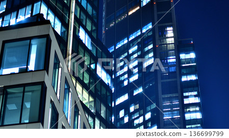 Big glowing windows in modern office building at night, in rows of windows light shines. Fragment of the glass facade of a modern corporate building at night. Modern glass office  in city. 136699799