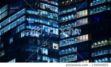 Big glowing windows in modern office building at night, in rows of windows light shines. Fragment of the glass facade of a modern corporate building at night. Modern glass office in city. Big glowing windows in modern office building at night, in rows of windows light shines. Fragment of the glass facade of a modern corporate building at night. Modern glass office in city. 136699805