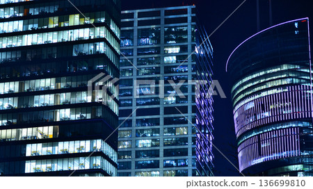 Big glowing windows in modern office building at night, in rows of windows light shines. Fragment of the glass facade of a modern corporate building at night. Modern glass office in city. Big glowing windows in modern office building at night, in rows of windows light shines. Fragment of the glass facade of a modern corporate building at night. Modern glass office in city. 136699810