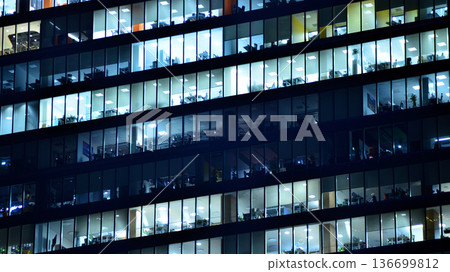 Big glowing windows in modern office building at night, in rows of windows light shines. Fragment of the glass facade of a modern corporate building at night. Modern glass office in city. Big glowing windows in modern office building at night, in rows of windows light shines. Fragment of the glass facade of a modern corporate building at night. Modern glass office in city. 136699812