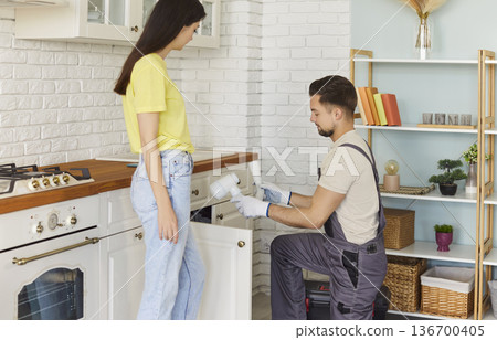 Man plumber in uniform cleaning siphon under kitchen sink, telling about problem to woman client 136700405