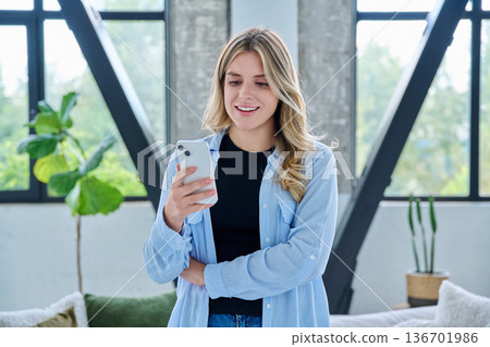 Young woman standing at home using smartphone Young woman standing at home using smartphone 136701986