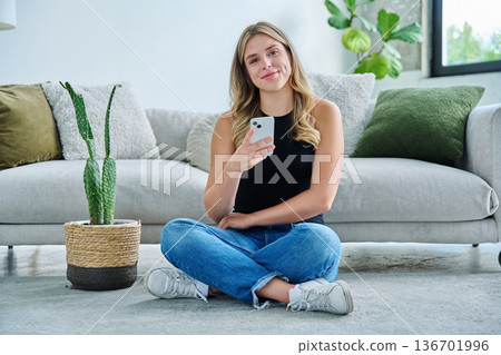 Smiling young woman with smartphone looking at camera sitting at home 136701996
