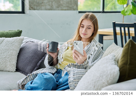 Relaxed beautiful young woman sitting at home on sofa with cup, using smartphone under blanket Relaxed beautiful young woman sitting at home on sofa with cup, using smartphone under blanket 136702014
