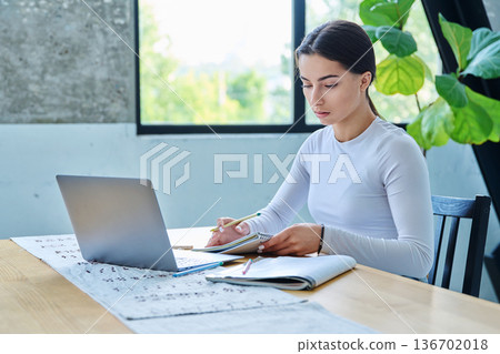 Teenage girl student sitting at the table with laptop, textbooks in home interior 136702018