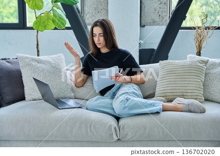 Young student girl study online, sitting at home on couch with laptop, textbook 136702020