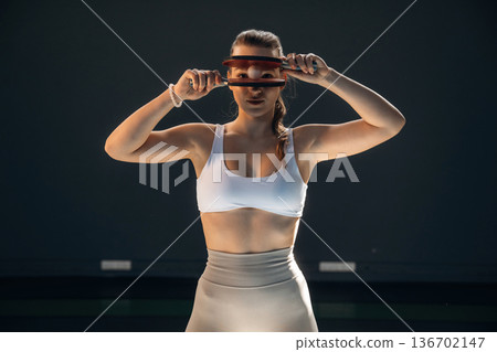 Holding ball between rackets. Beautiful young woman is by the tennis table indoors Holding ball between rackets. Beautiful young woman is by the tennis table indoors 136702147