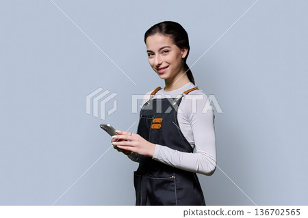 Portrait of young smiling woman in apron holding smartphone on gray background 136702565