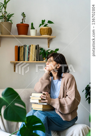 Mature woman daydreaming holding books at home Mature woman daydreaming holding books at home 136702804