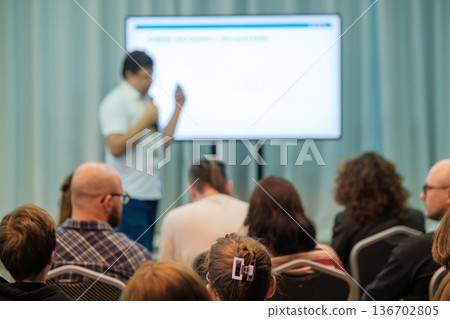 Presenter delivers lecture to engaged audience in conference room with large screen display during event. 136702805